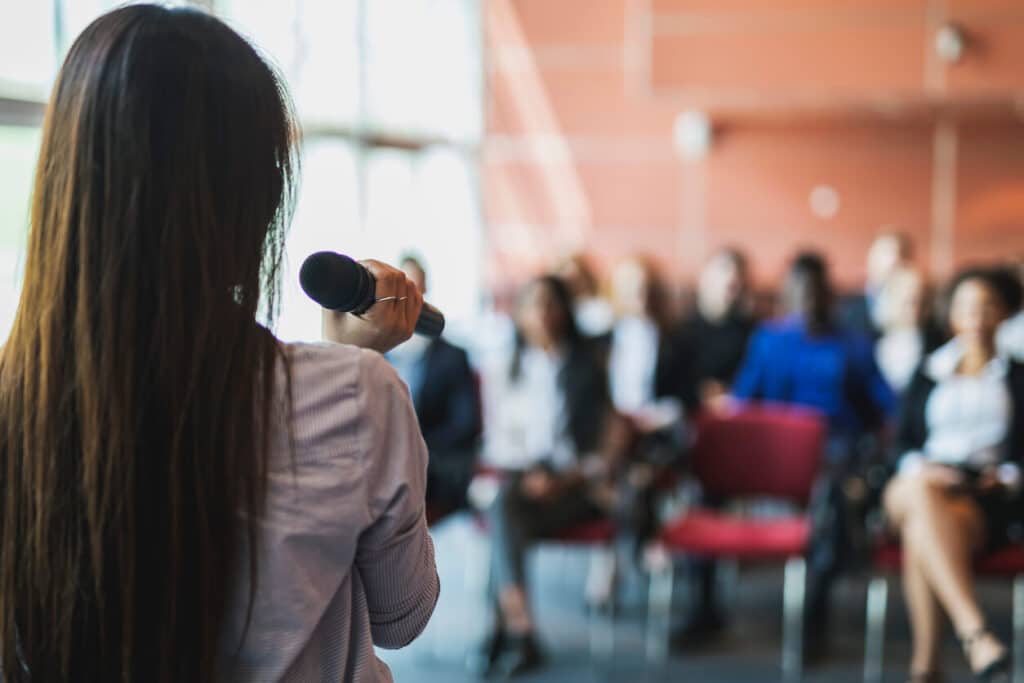 A female presenter giving a speech to a small audience.